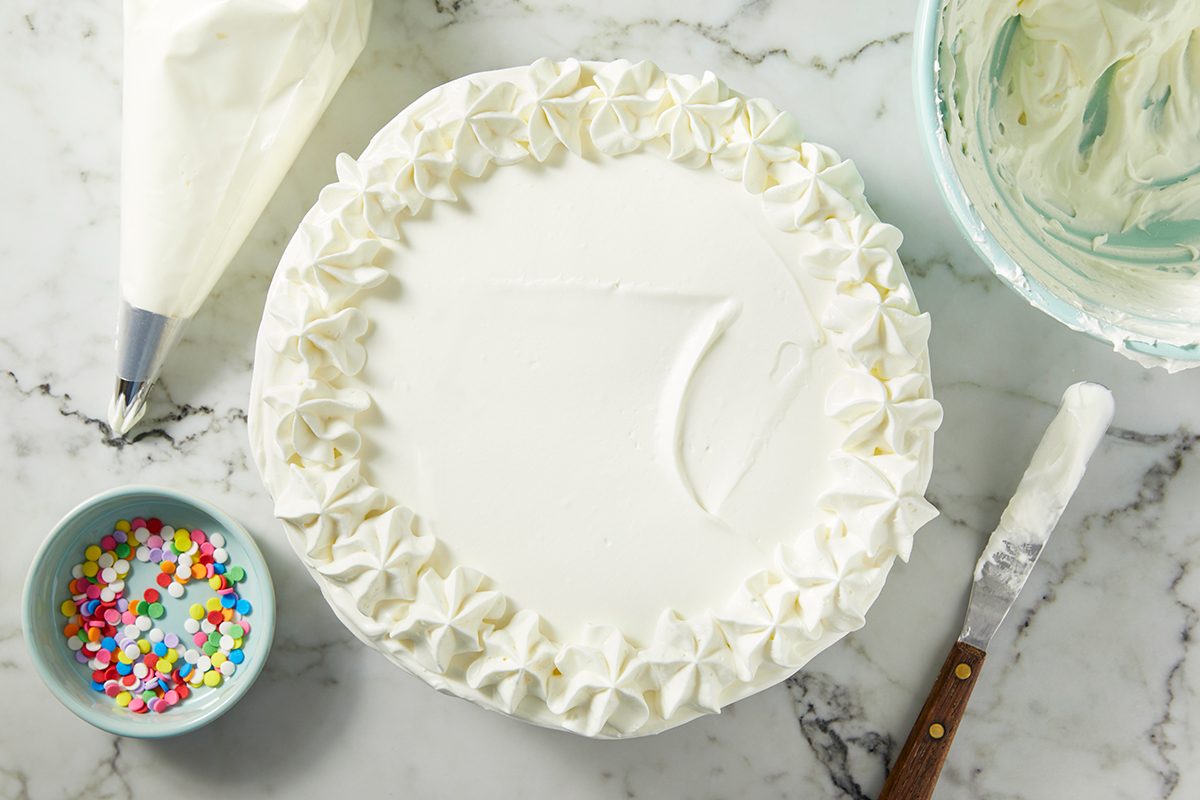 A white frosted cake on a marble surface, decorated with piped cream swirls around the edge. 