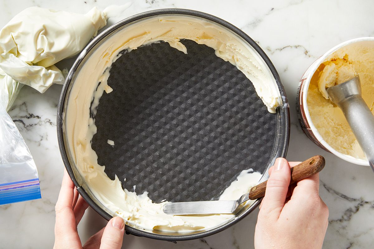 A person uses an offset spatula to spread softened ice cream into a round springform pan on a marble countertop. 