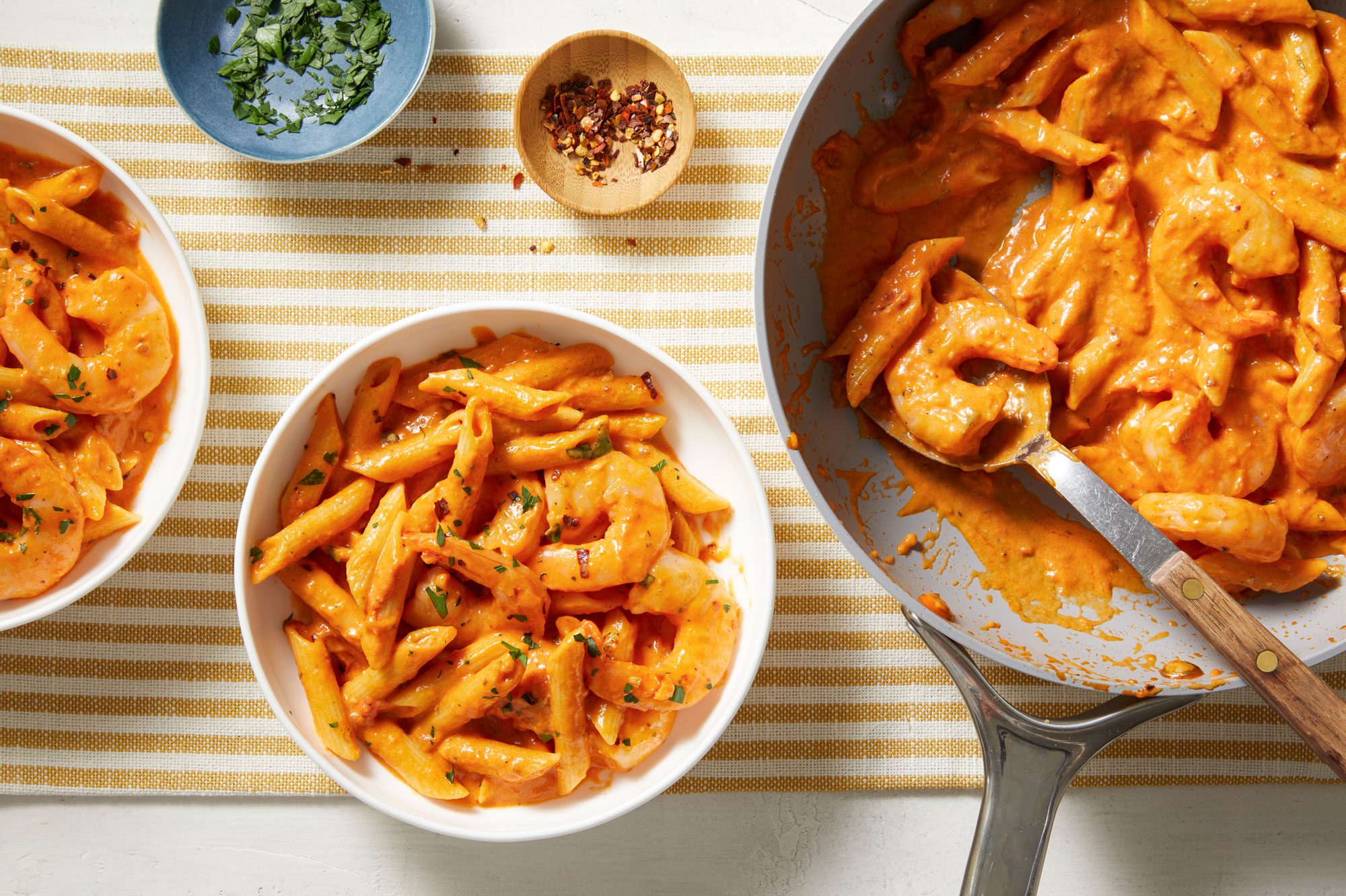 Overhead shot of Creamy Tomato Shrimp with Penne; in a skillet with a serving spoon; served in two bowls; sprinkle with parsley and chilli flakes; all set on a napkin; white surface