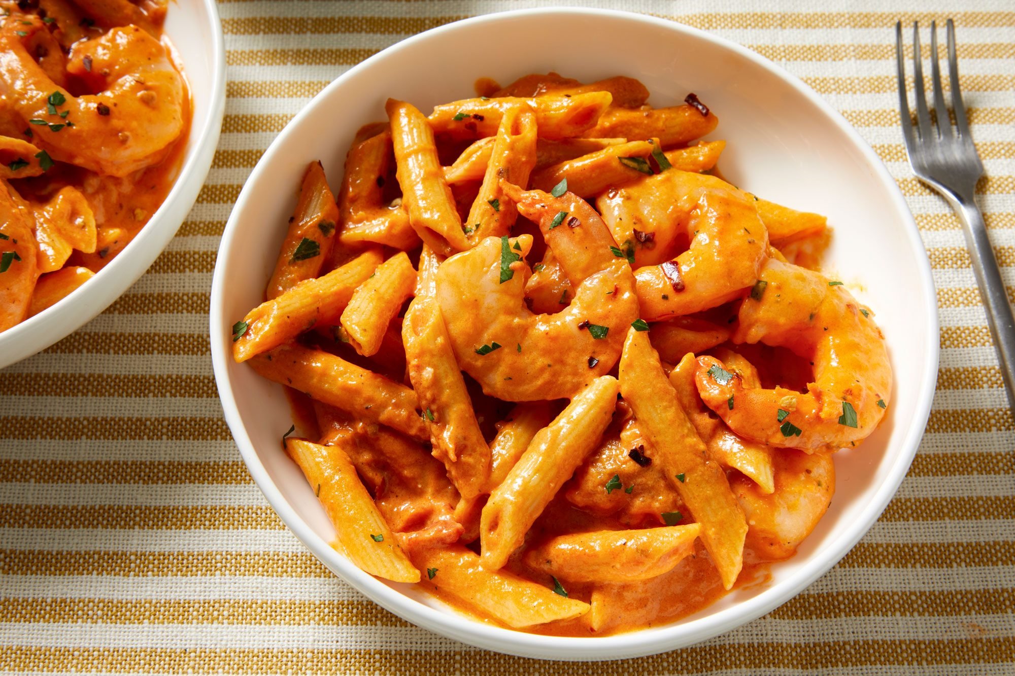 Close-up shot of Creamy Tomato Shrimp with Penne; served in two bowls with a fork; sprinkle with parsley and chilli flakes; all set on a napkin