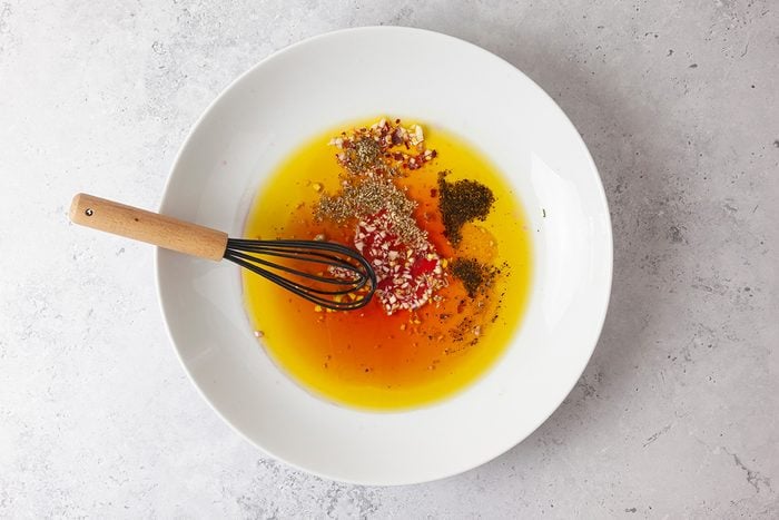 A white bowl holds oil, herbs, and seasonings being mixed with a whisk on a light, textured surface.