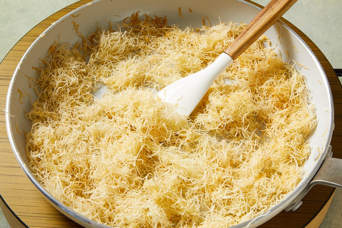 A white frying pan filled with lightly toasted shredded phyllo dough, being stirred with a white spatula that has a wooden handle.