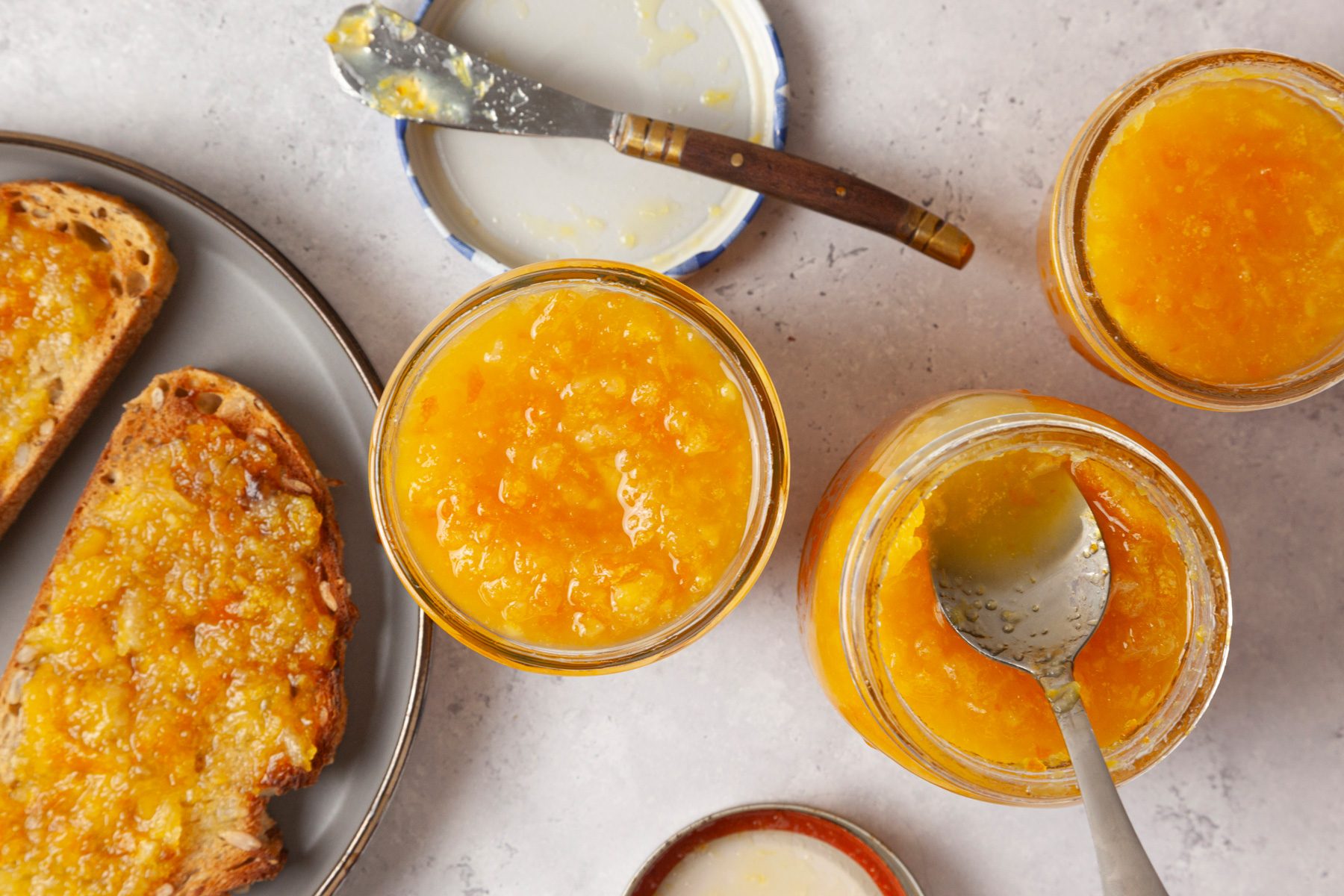 overhead shot of spread of orange marmalade presented in clear glass jars, two slices of toasted bread, topped with the marmalade, rest on a round gray plate; one jar is slightly open, revealing a spoon resting inside it, while the others sit upright, a butter knife, partially covered in marmalade, lies beside an empty lid