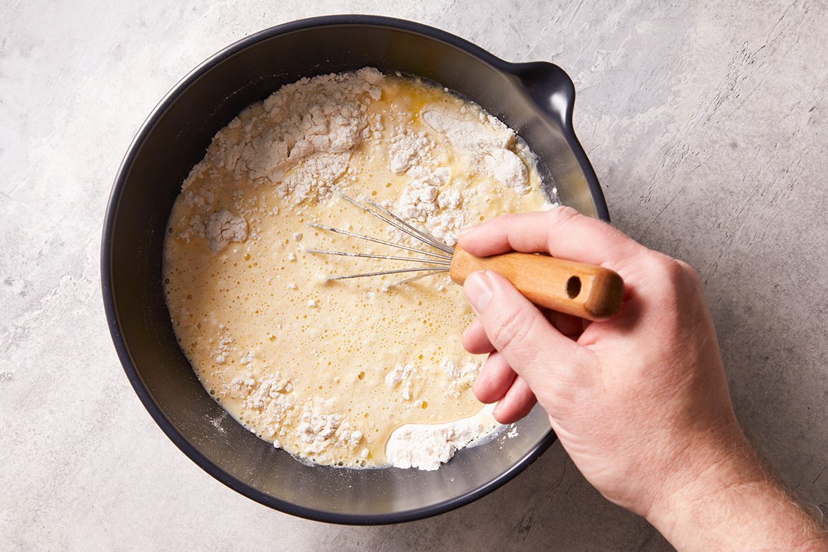 A hand holding a whisk mixes flour and liquid ingredients in a black bowl on a light countertop, preparing a batter or dough.