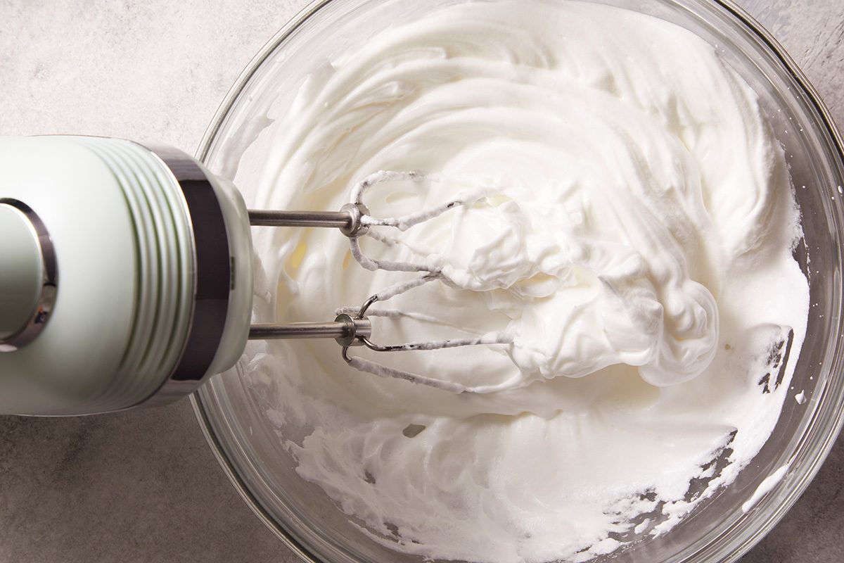 A hand mixer with metal beaters is whipping fluffy, white cream or egg whites in a clear glass bowl on a light-colored surface.