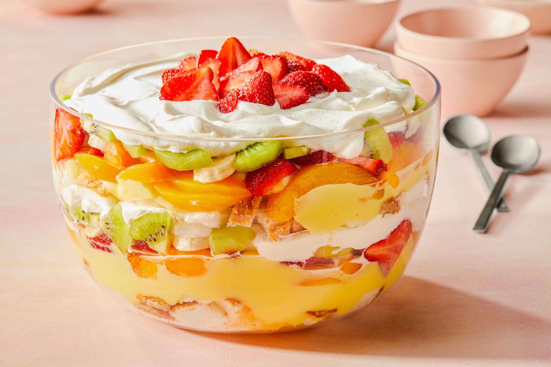 Horizontal AP; Pull shot of Fruit Trifle in a large, transparent bowl; In the background, there are soft pink bowls and silver spoons