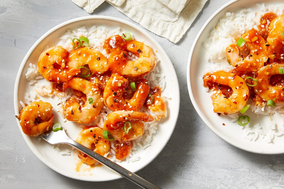 Two white bowls filled with white rice topped with glazed shrimp, garnished with sliced green onions and black sesame seeds, with a fork resting in one bowl on a light surface.