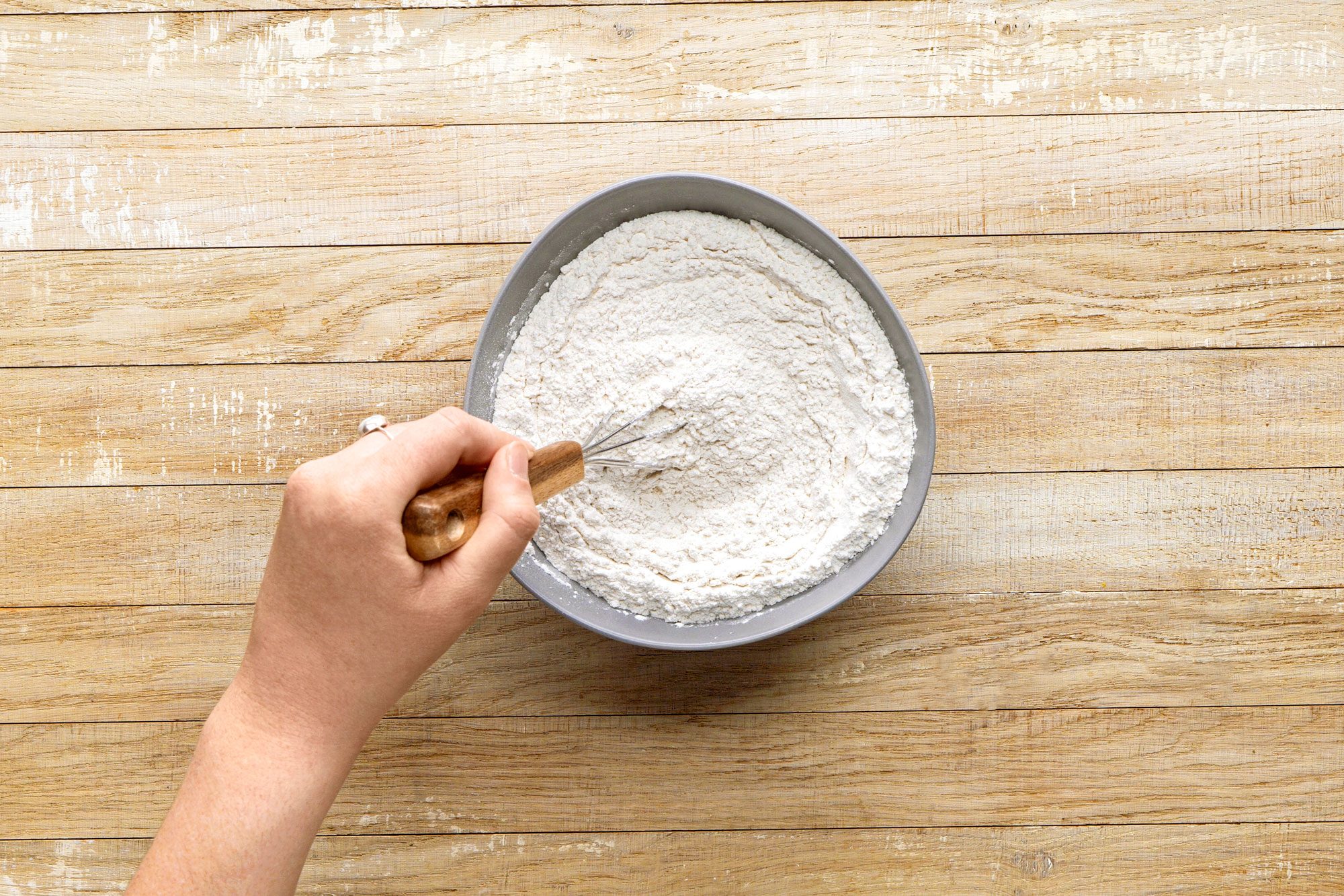 overhead shot of a hand stirring a bowl of flour with a whisk; the bowl is circular and gray, the background is made of wooden planks