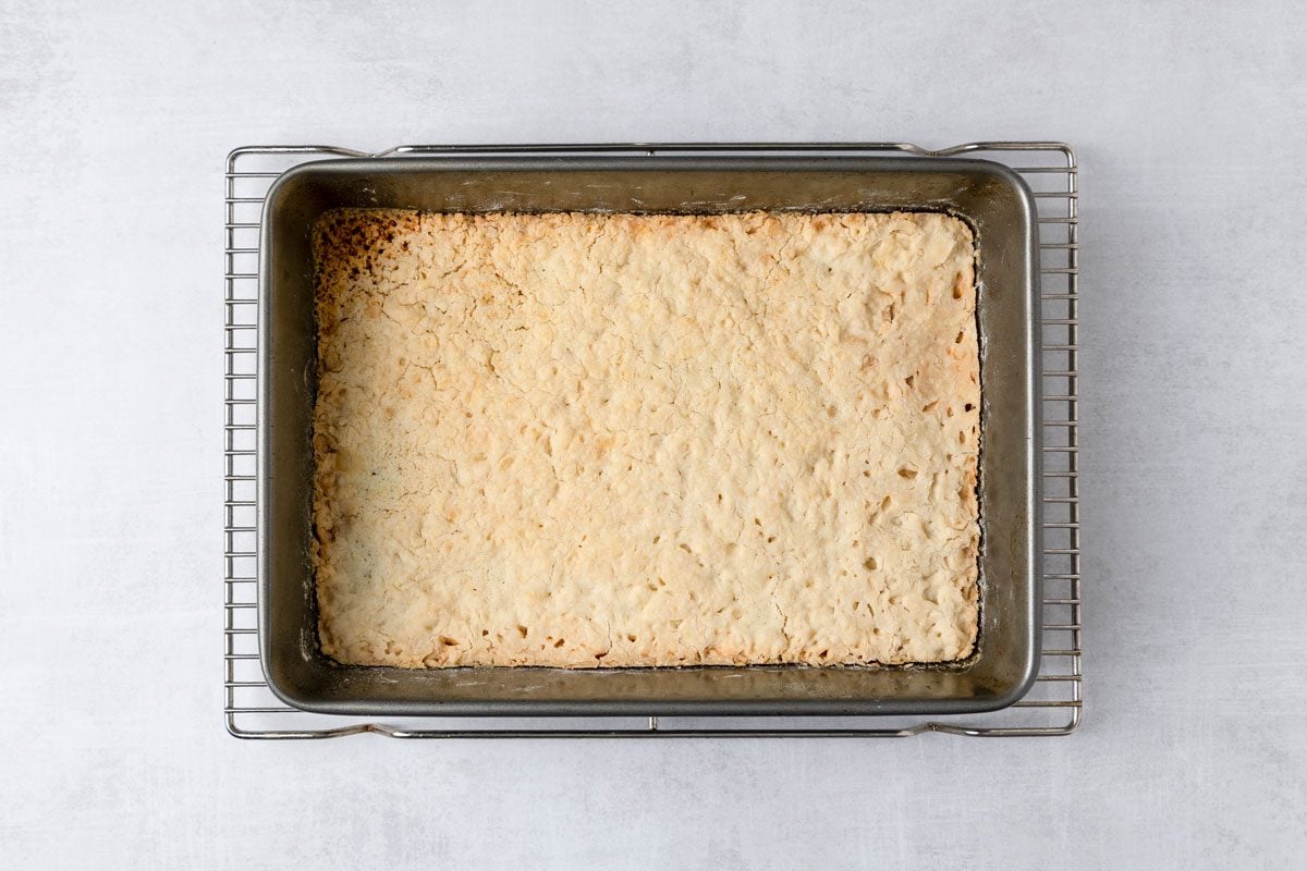overhead shot of a baking pan placed on a cooling rack; the pan contains a large slab of baked goods, the pan itself is metallic and rectangular, while the surface beneath is smooth and light colored