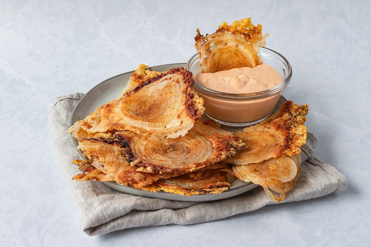 Plate of crispy, golden artichoke chips served with a bowl of dipping sauce on a soft, neutral-toned napkin, set against a light gray background.