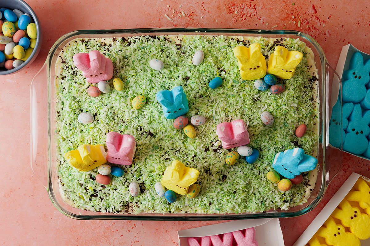 Birds eye view of easter dirt cake in a glass baking dish with green coconut grass, chocolate eggs, and pastel marshmallow rabbits with candy packaging on the sides