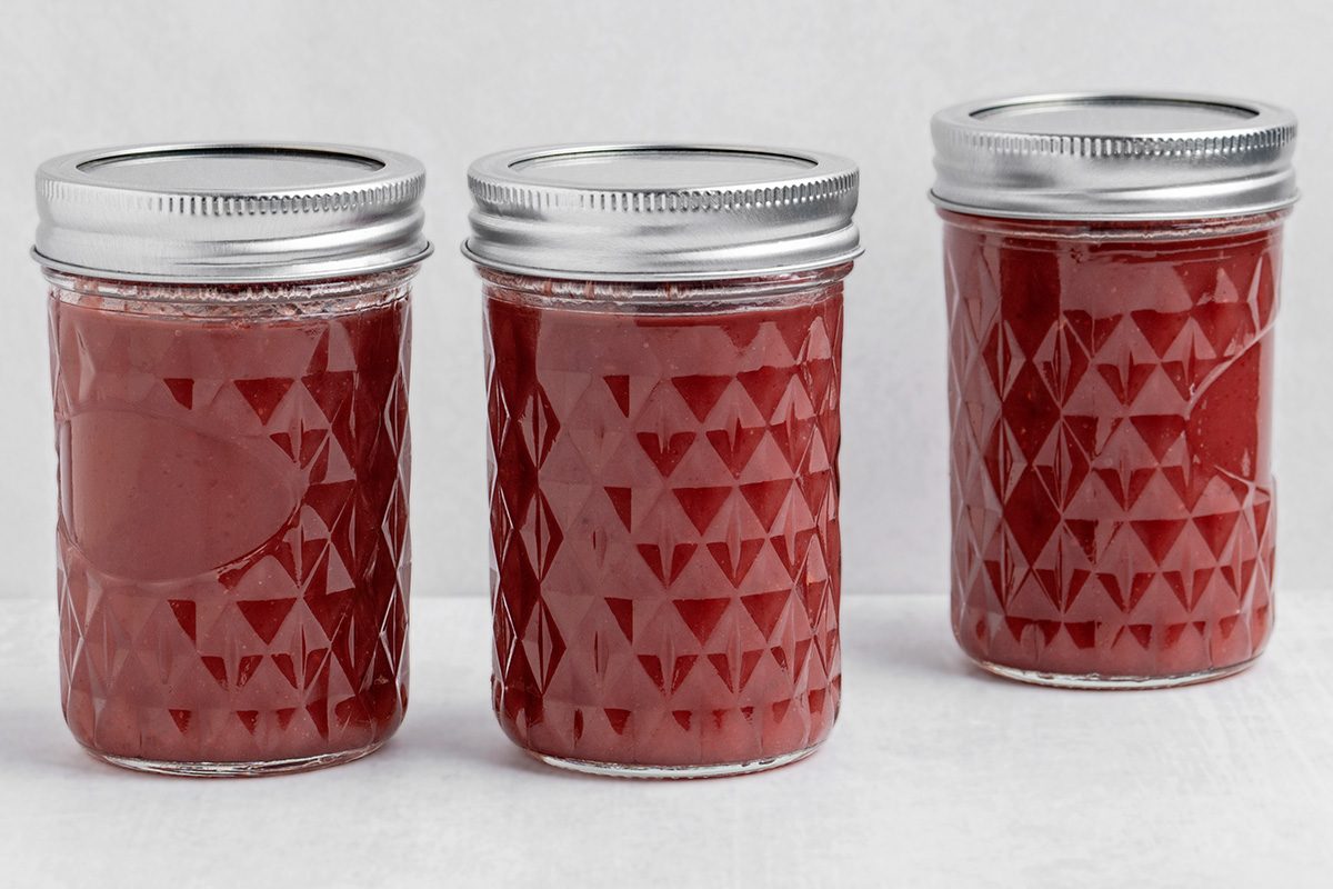 Three glass jars with silver lids are filled with red jam or preserves and arranged in a row against a plain white background. The jars have a textured geometric pattern.