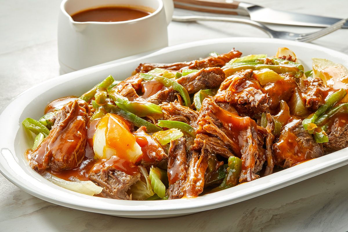 A white platter filled with shredded beef, green beans, onions, and brown gravy, with a small white jug of extra sauce in the background, and utensils on a marble surface.