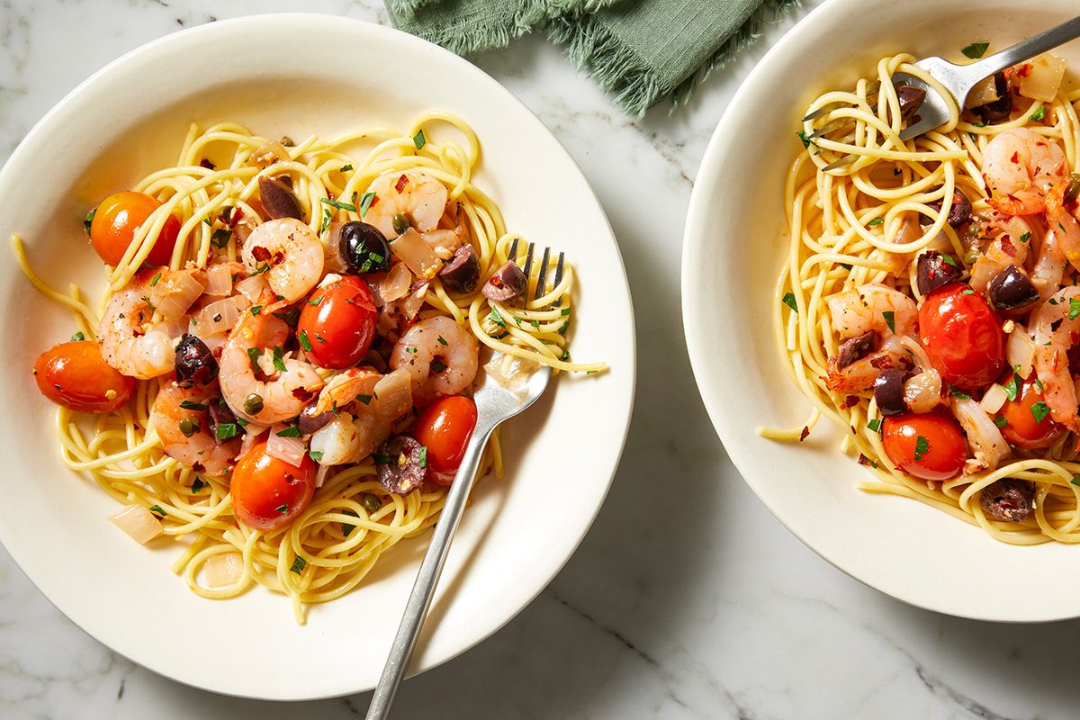 Two bowls of pasta with shrimp, cherry tomatoes, olives, and herbs. Each dish is garnished with pepper flakes and served with a fork. The bowls are on a marble surface with a green cloth in the background.