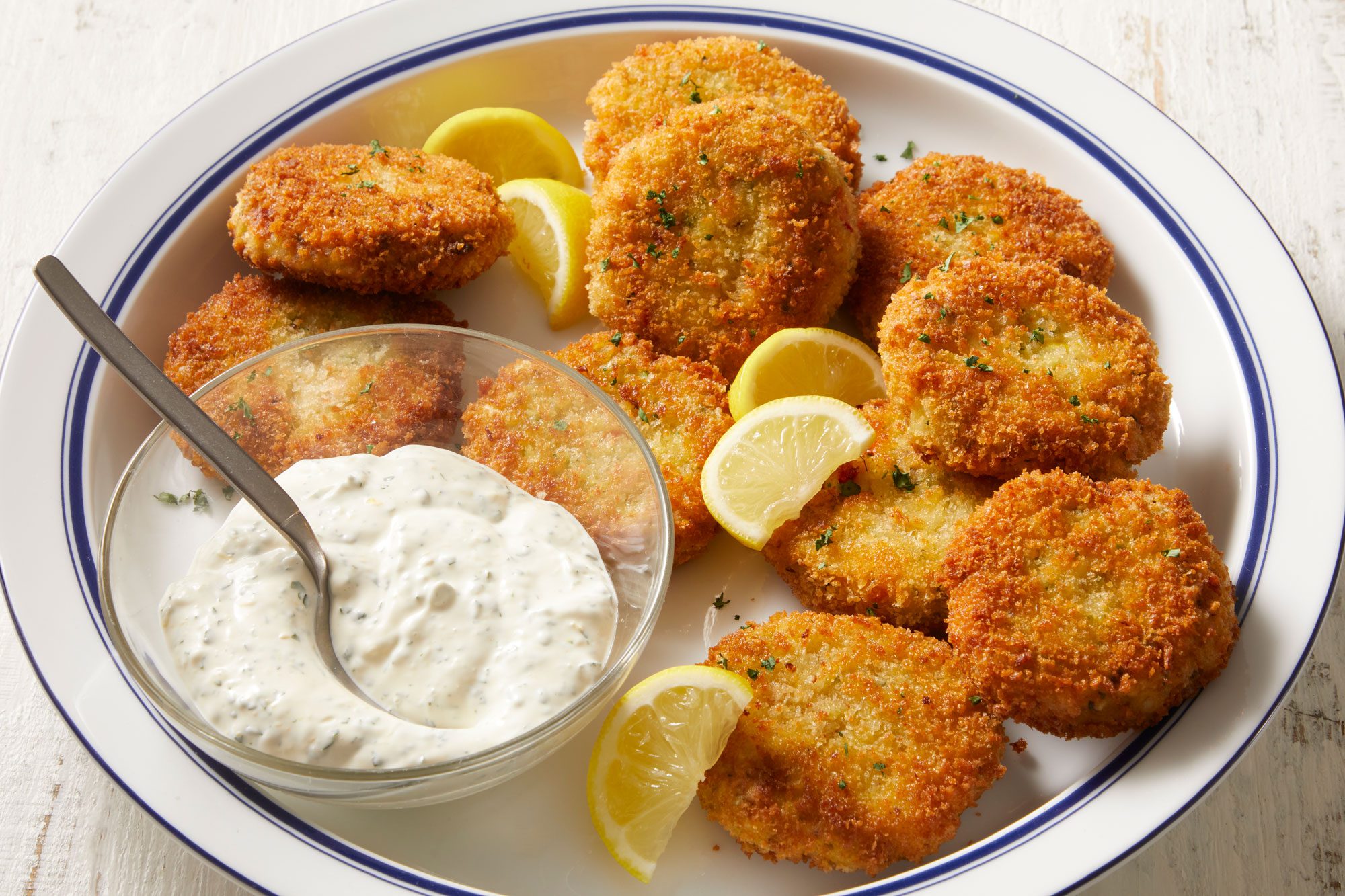 3/4th shot of a plate of Company Shrimp and Crab Cakes arranged neatly, and accompanied by fresh lemon wedges; beside them lies a bowl of creamy sauce with a spoon, the background is a simple white wooden surface