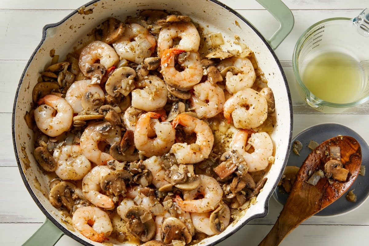Overhead shot of add the shrimp; broth and seasonings; Bring to a boil; Reduce heat; cook and stir for 4-5 minutes or until sauce is slightly thickened and shrimp turn pink; wooden spoon; white wooden surface;