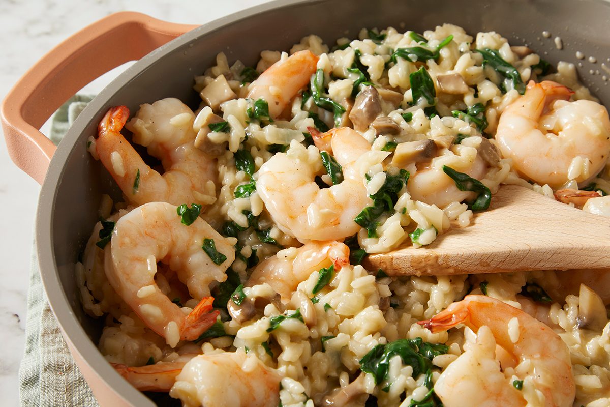 A pot of shrimp risotto with spinach and mushrooms. A wooden spatula is mixing the creamy rice dish, with shrimp and greens visible throughout.