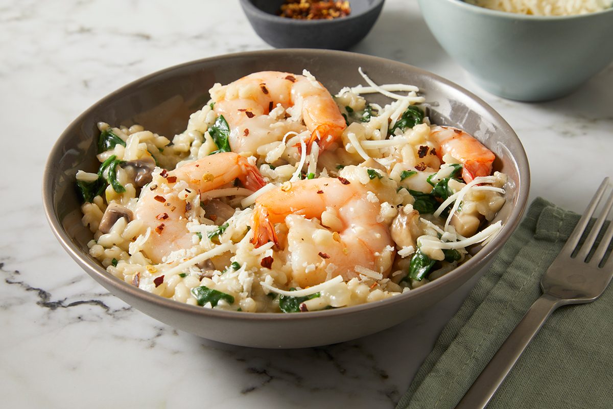 A bowl of pasta with shrimp, spinach, and grated cheese on a marble surface. A small bowl of red pepper flakes and another bowl of grated cheese are in the background. A fork and green napkin are placed beside the bowl.