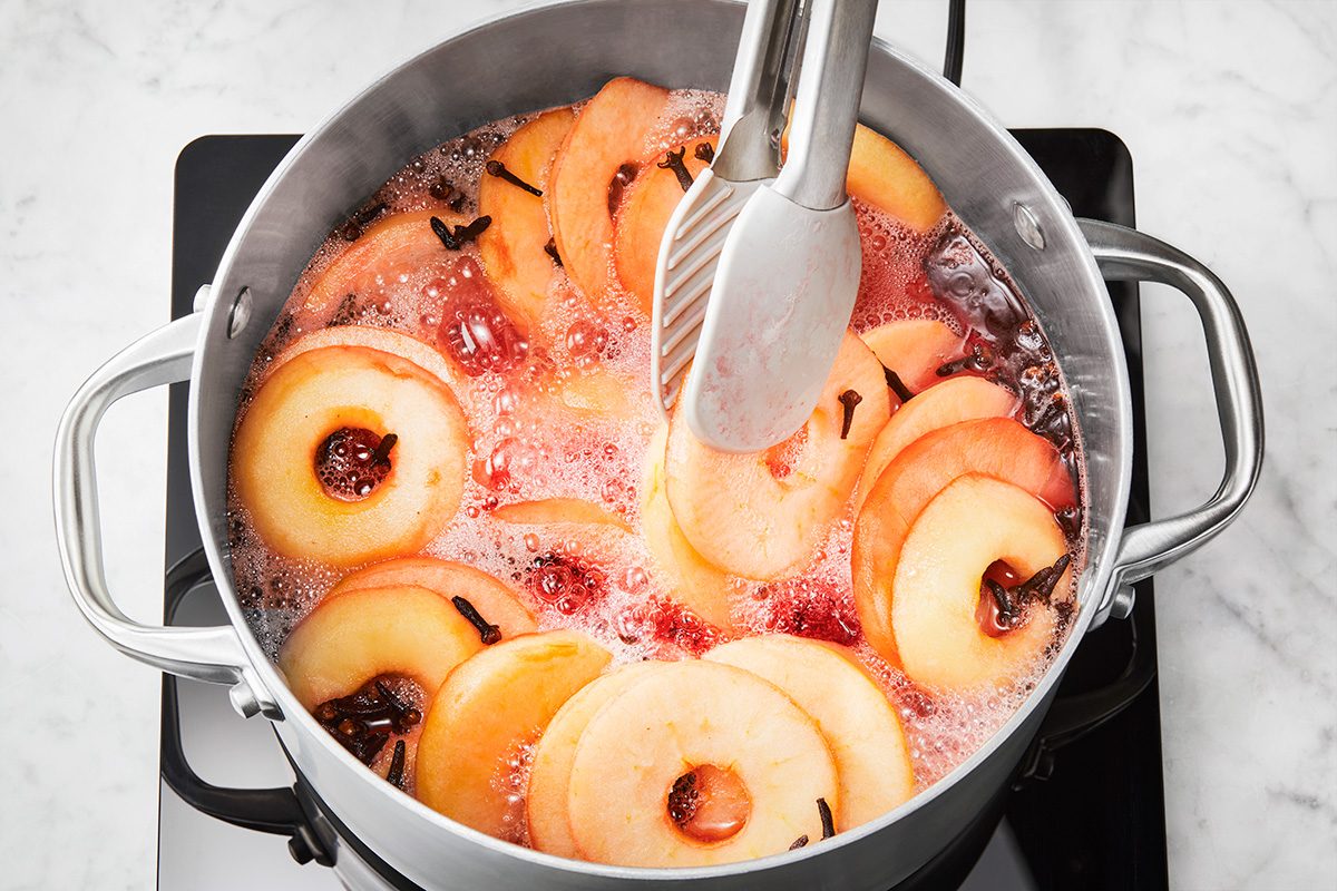 Sliced apples with cloves simmer in a pot of bubbling liquid on a stovetop, with metal tongs resting inside the pot.