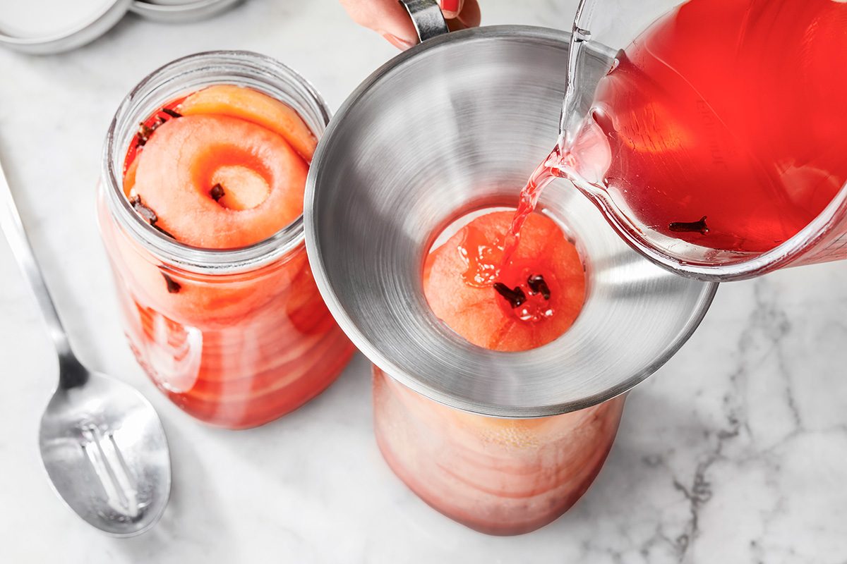 A metal funnel sits on a glass jar filled with whole apples and red liquid, while more red liquid is being poured in from a pitcher. Another jar with apples and red liquid and a spoon are nearby on a marble surface.