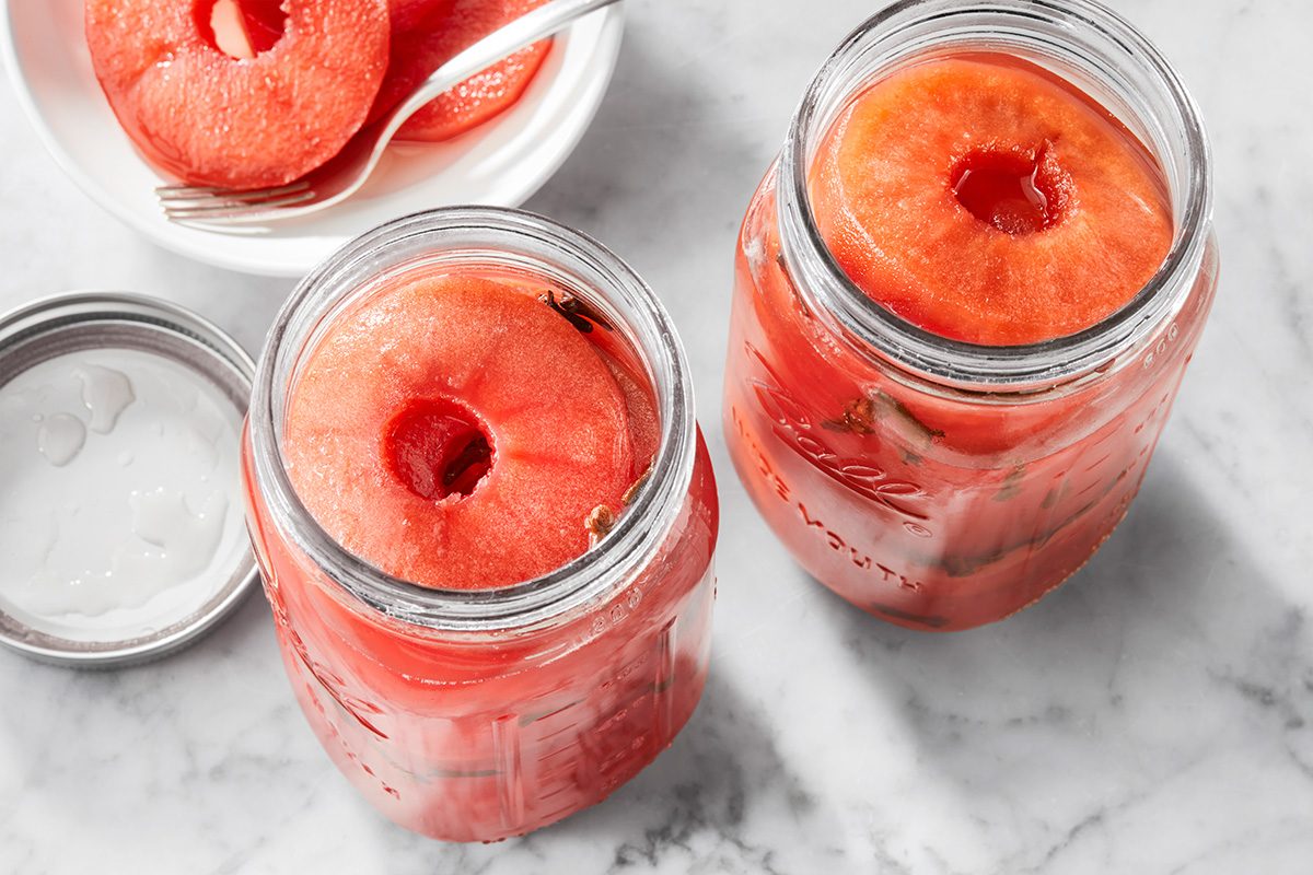 Two glass jars filled with large, round slices of pickled watermelon rind sit on a marble surface. A lid is beside one jar, and a plate with more watermelon slices and a fork is in the background.