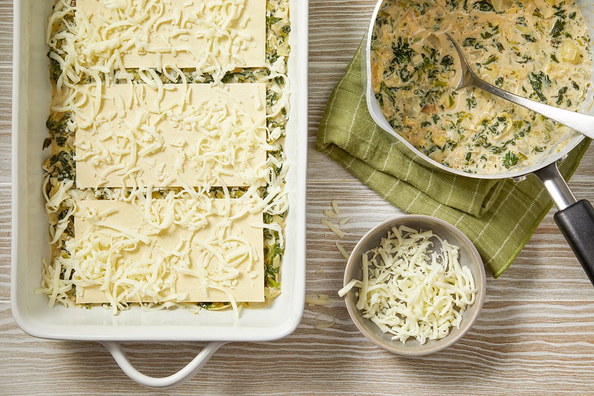 A white baking dish with layered lasagna sheets and grated cheese. Beside it, a bowl of cheese and a saucepan with creamy spinach sauce rest on a green cloth. All items are on a light wood table.
