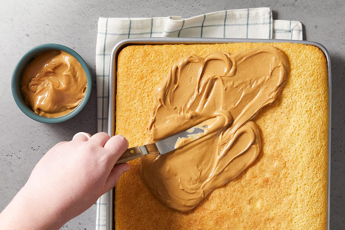 A hand spreading creamy peanut butter over a square, golden-brown cake with a spatula. A bowl of peanut butter sits nearby on a checked cloth.