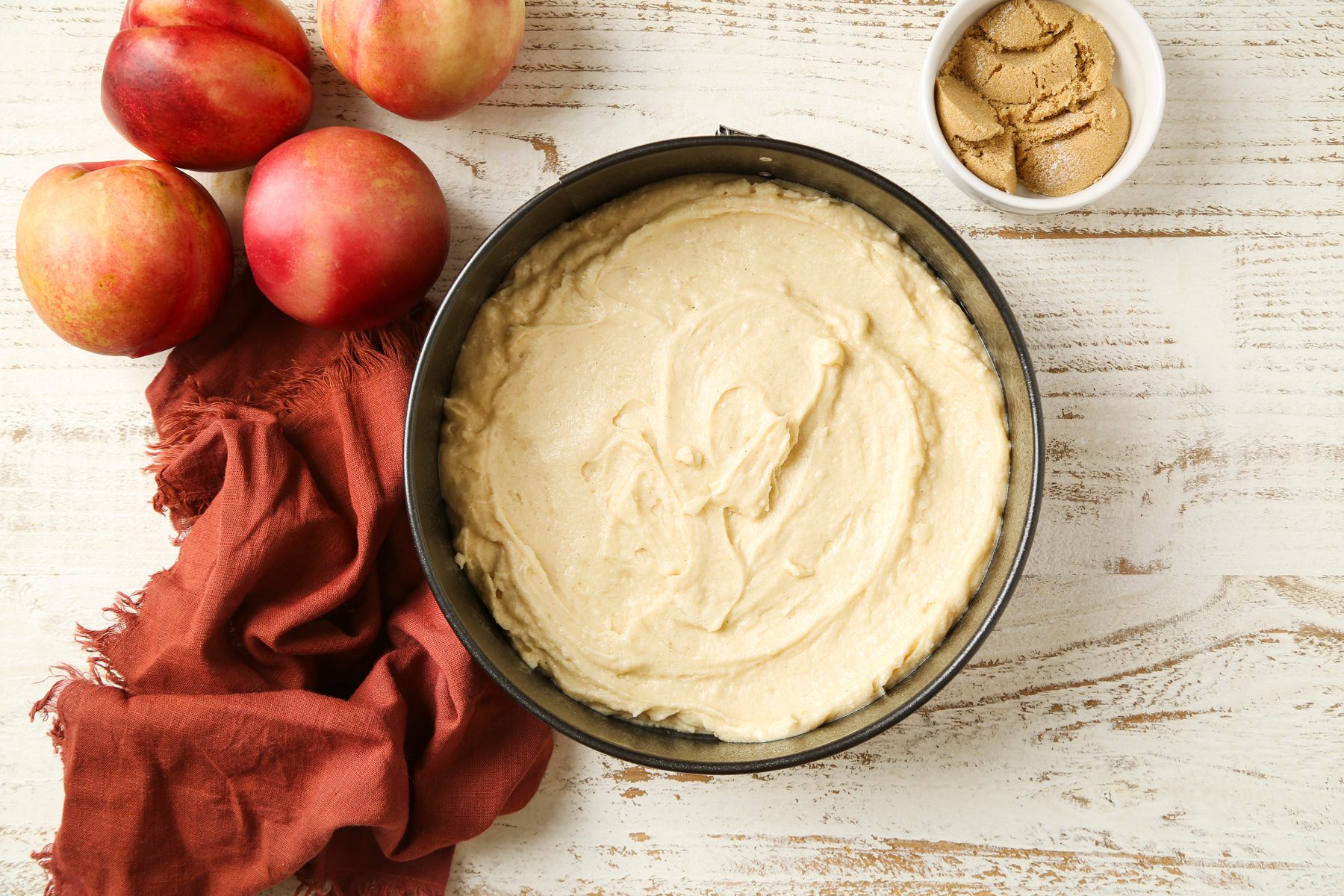 Overhead shot; Preheat oven to 350 degree; In a large bowl cream sugar and butter until light and fluffy 5-7 minutes; Beat in egg; vanilla extract and almond extract; Combine flour; baking powder; cinnamon and salt; add to creamed mixture alternately with buttermilk; beating well after each addition; Pour batter into a greased 9 inch springform pan; peaches; a napkin; white wooden surface;
