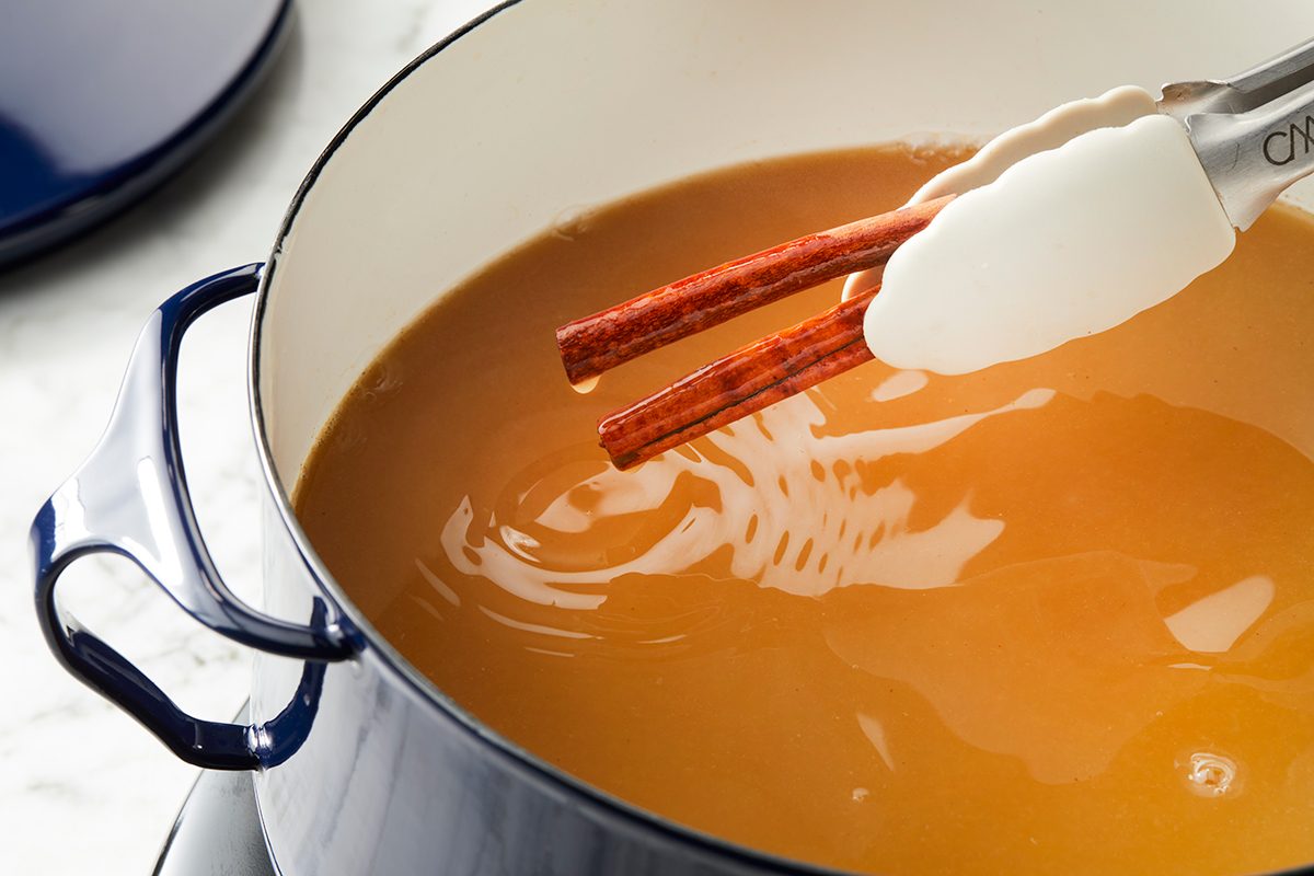A pair of tongs holds two cinnamon sticks over a pot filled with brown liquid, possibly a beverage or broth, on a stovetop.