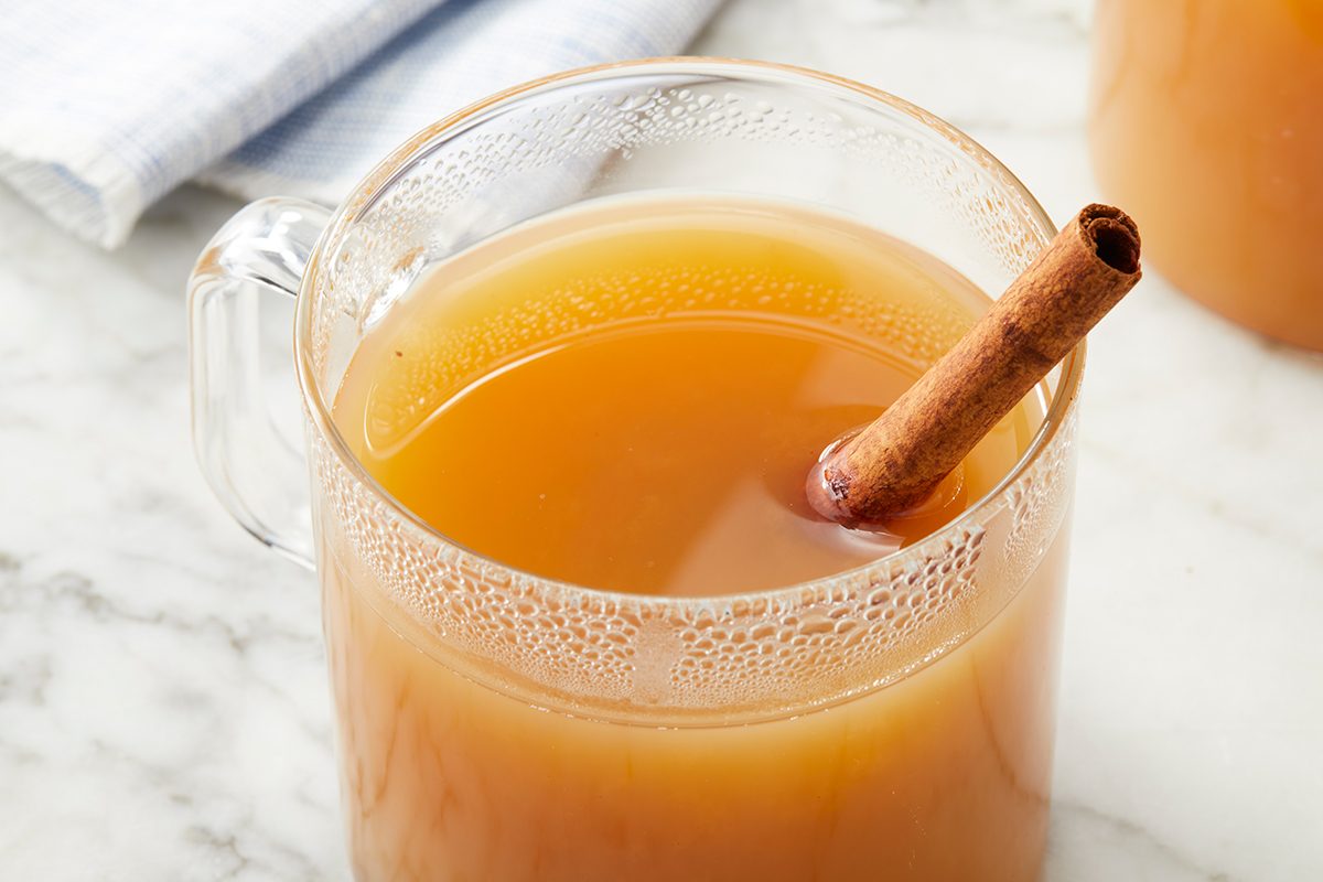 A clear glass mug filled with warm apple cider, garnished with a cinnamon stick, sits on a marble surface. A folded white cloth is partially visible in the background.