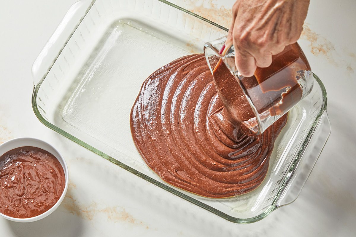 Overhead shot: a hand is seen spreading the mixture into a greased clear glass baking dish using a spatula; set aside; Next to the baking dish, a small white bowl filled with a similar brown mixture is visible; All items are set on a marble surface;
