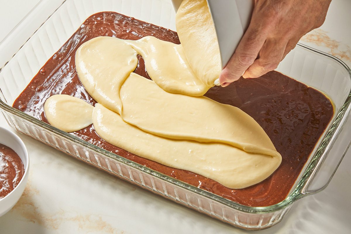 High angle shot of a glass baking sheet; In a small bowl beat the cream cheese; butter; sugar; flour and vanilla until smooth; add eggs; beat on low speed just until combined; Spread evenly over brownie batter; all set on a marble surface;