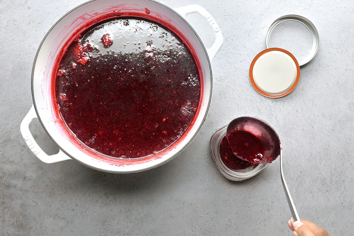 A pot filled with homemade red jam sits on a gray surface. A hand is spooning some jam into a glass jar, with a jar lid placed nearby.