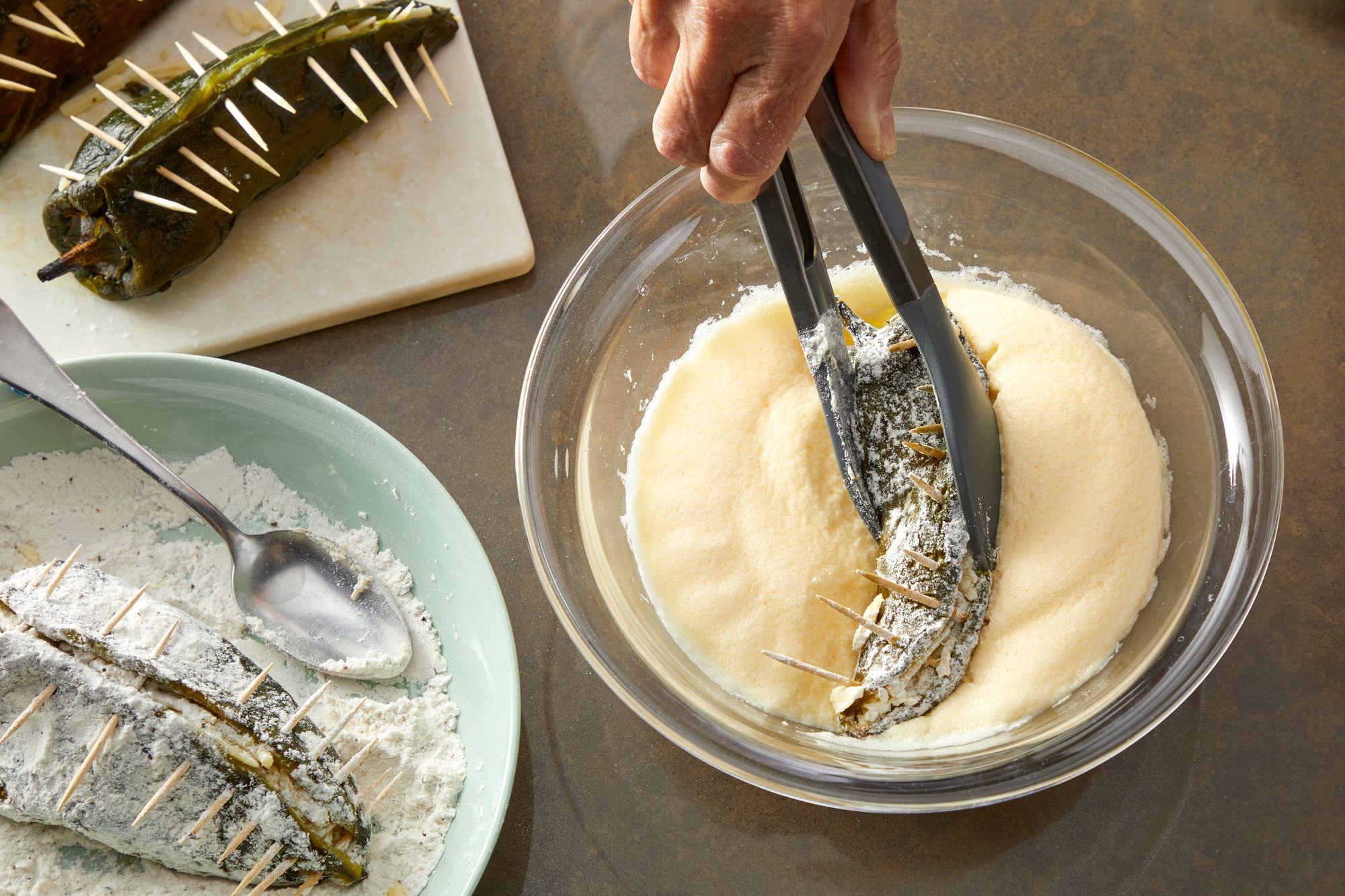 Overhead shot of place flour in a shallow bowl; stir in salt and pepper; Dip stuffed peppers in the flour mixture; then egg mixture with use of tongs; Place in hot oil; all arranged on a light brown surface