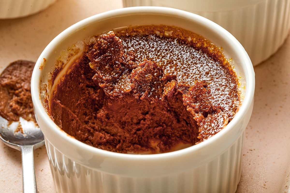 A chocolate soufflé in a white ramekin, partially eaten to reveal its moist, airy texture. A dusting of powdered sugar tops the dessert, and a spoon rests nearby on the table.