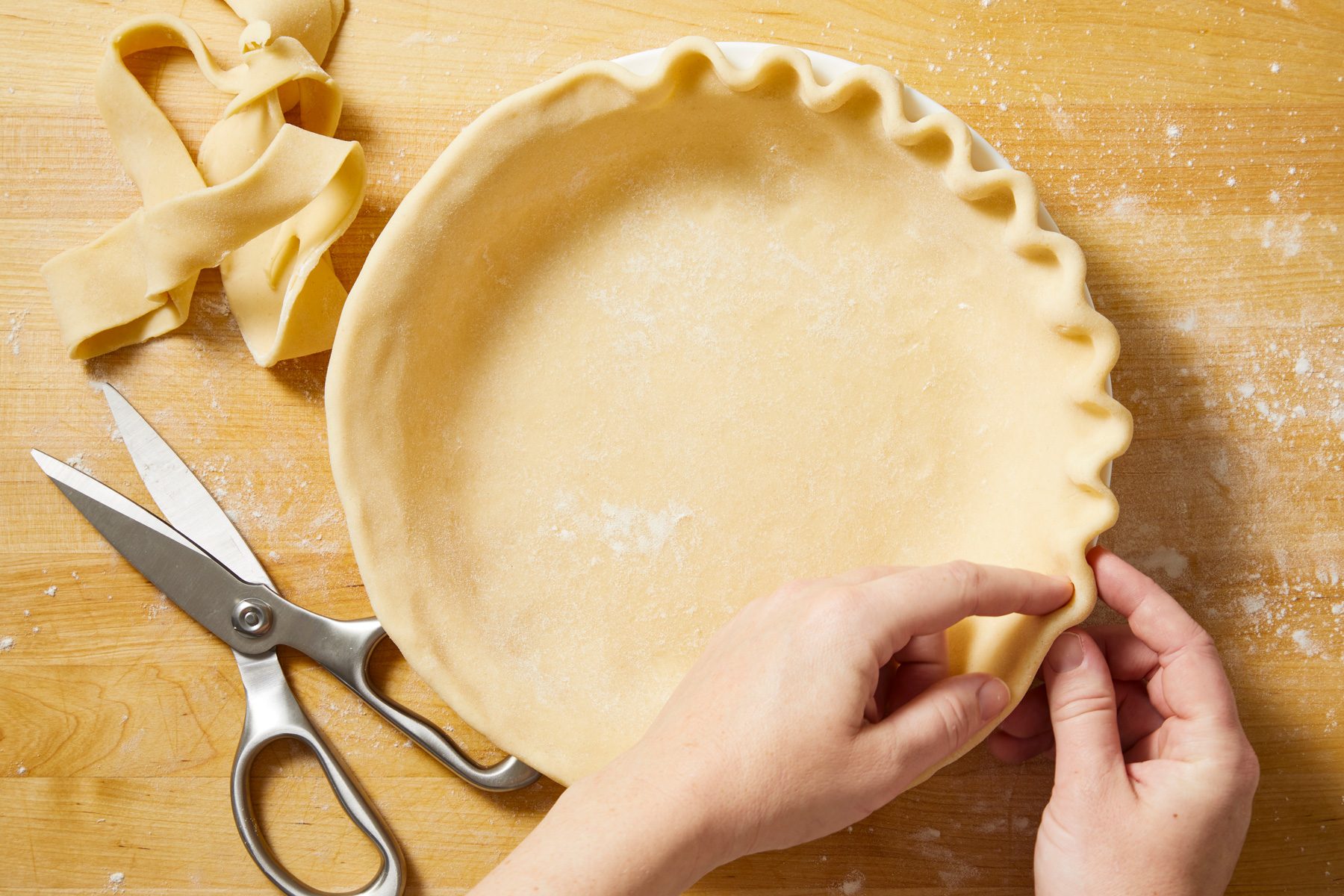 overhead shot of a hand preparing pie crust resting in a pie dish; the crust has a fluted edge, next to the pie dish, there are some strips of dough, possibly scraps; a pair of silver scissors is positioned nearby on the wooden surface, the table is lightly dusted with flour