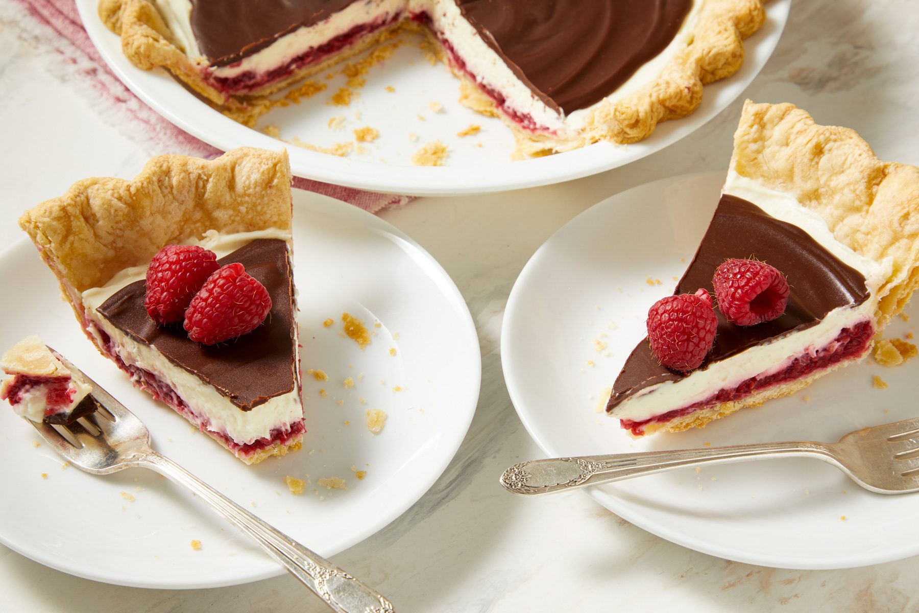 3/4th shot of two Chocolate Raspberry Pie slices; underneath the chocolate layer is a vibrant raspberry filling, which is visible in the sectioned slice; two slices of the pie are arranged on white plates, each garnished with fresh raspberries on top; a fork rests beside one of the slices