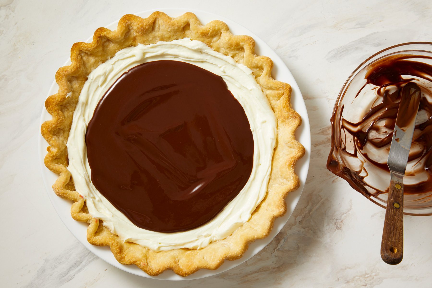overhead shot of a flaky, golden brown crust that is neatly arranged with a smooth layer of white cream filling around the edges; at the center, a glossy, rich layer of chocolate filling, beside the pie, there is a glass bowl with remnants of chocolate and cream mixture