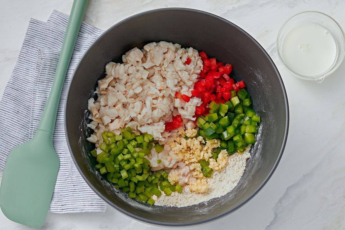 A mixing bowl with chopped shrimp, red and green bell peppers, celery, flour, and grated cheese, next to a green spatula, a striped cloth, and a small glass of milk on a white surface.