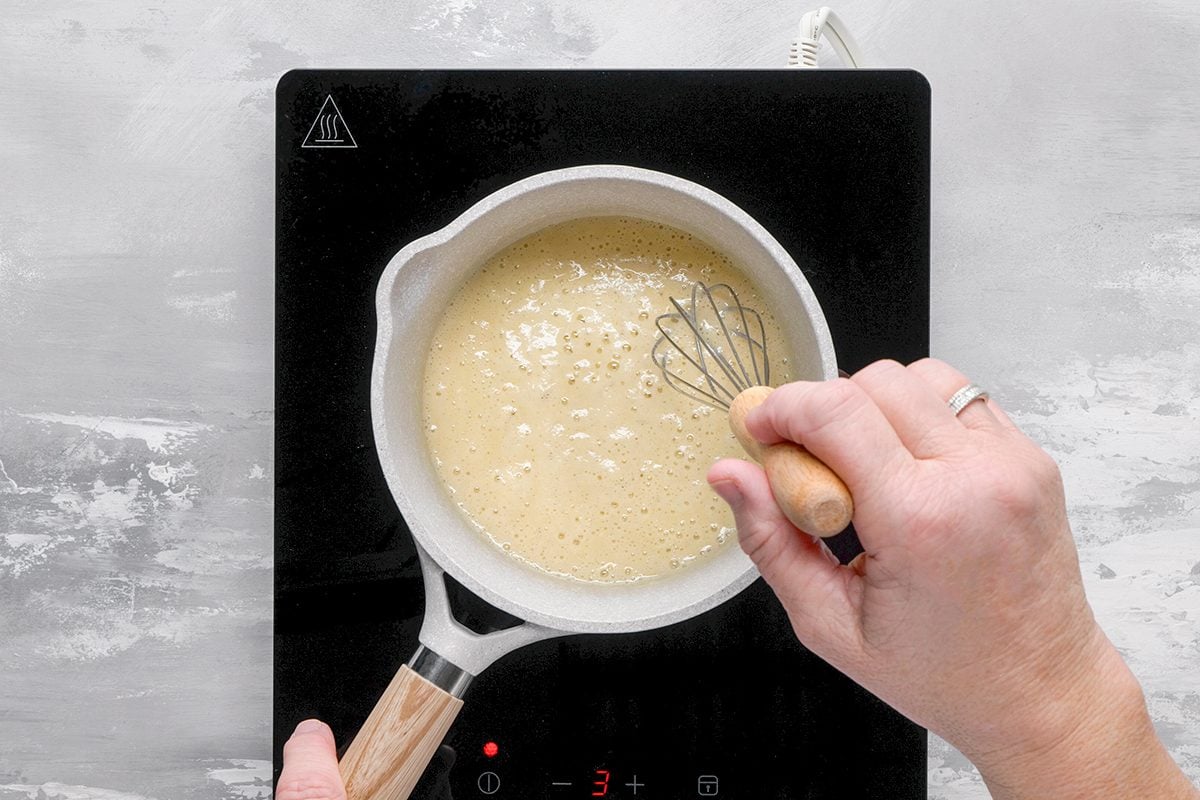 A person whisks a creamy mixture in a white saucepan on a black induction cooktop, viewed from above.