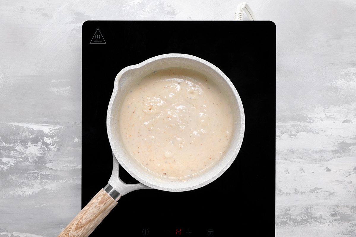 A white saucepan filled with a bubbly, light-colored mixture sits on a black electric stovetop against a light gray, textured background.