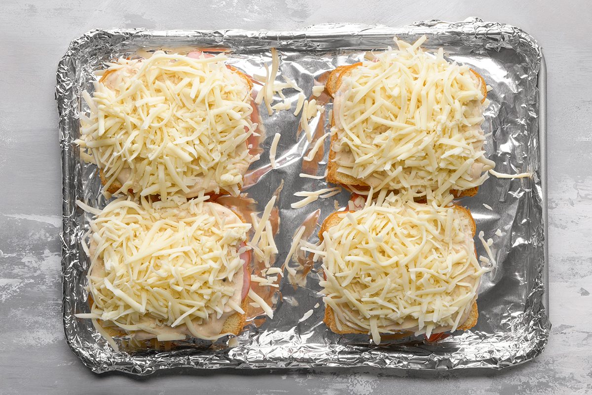 Four open-faced sandwiches topped with a generous amount of shredded cheese are arranged on a foil-lined baking sheet, ready to be baked.