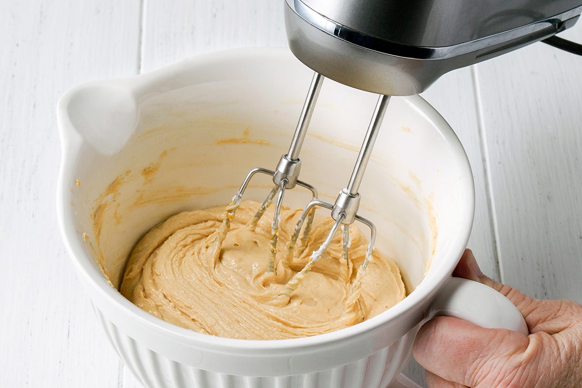 A hand holds a white mixing bowl while an electric hand mixer blends creamy batter inside the bowl on a white surface.
