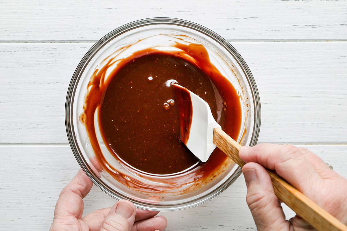 A person stirs chocolate batter in a clear glass bowl with a white spatula on a white wooden surface.