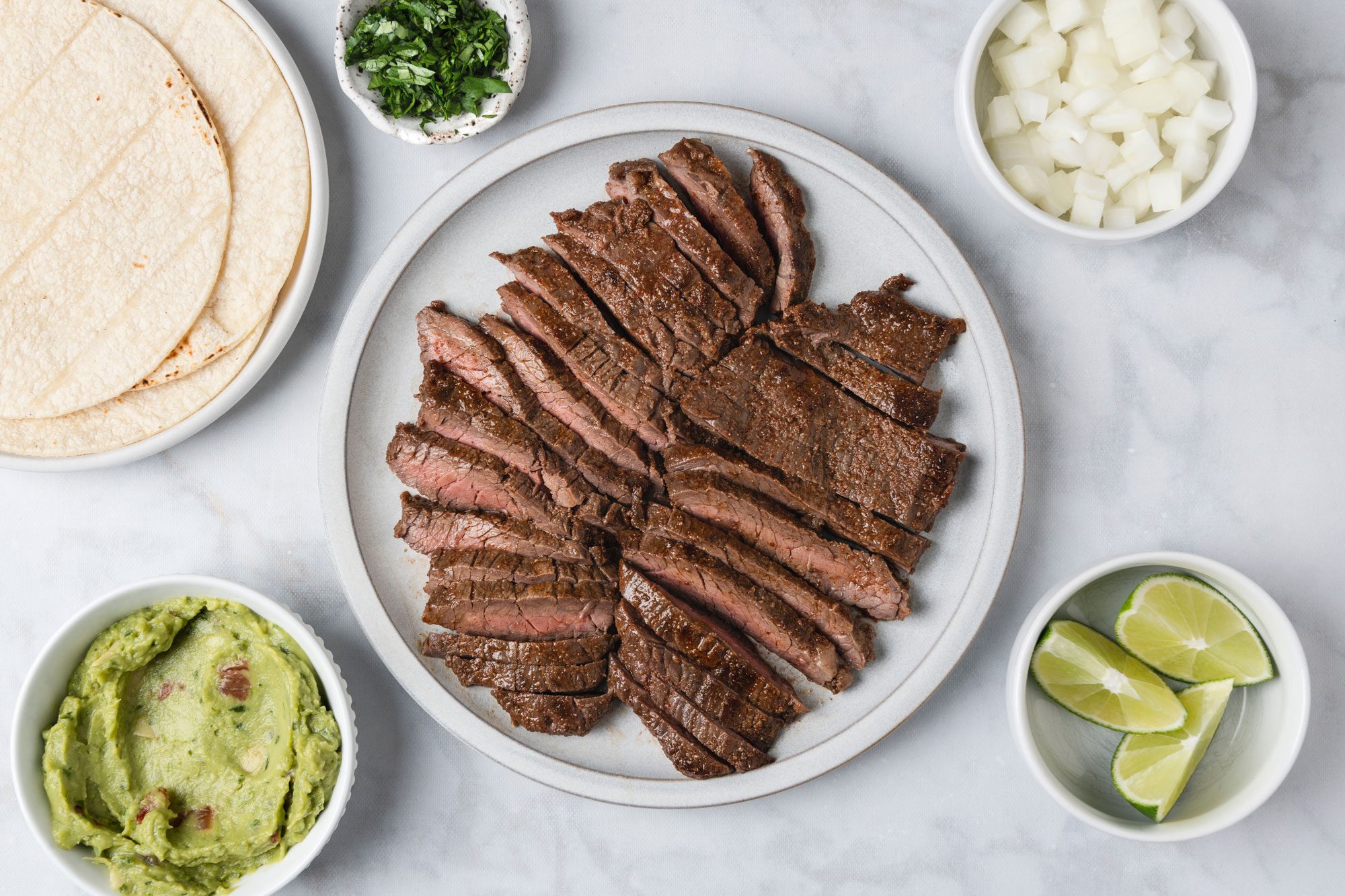 overhead shot of Sliced grilled steak on a plate, surrounded by bowls of chopped onions, lime wedges, guacamole, chopped cilantro, and a stack of tortillas on a light marble surface