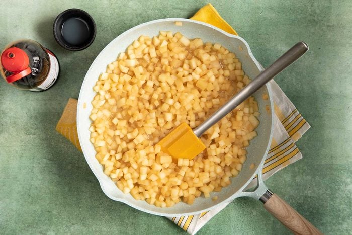 Overhead shot of cooking the apple filling in ceramic cookware kept on top of a green marble countertop, with maple syrup kept beside it.