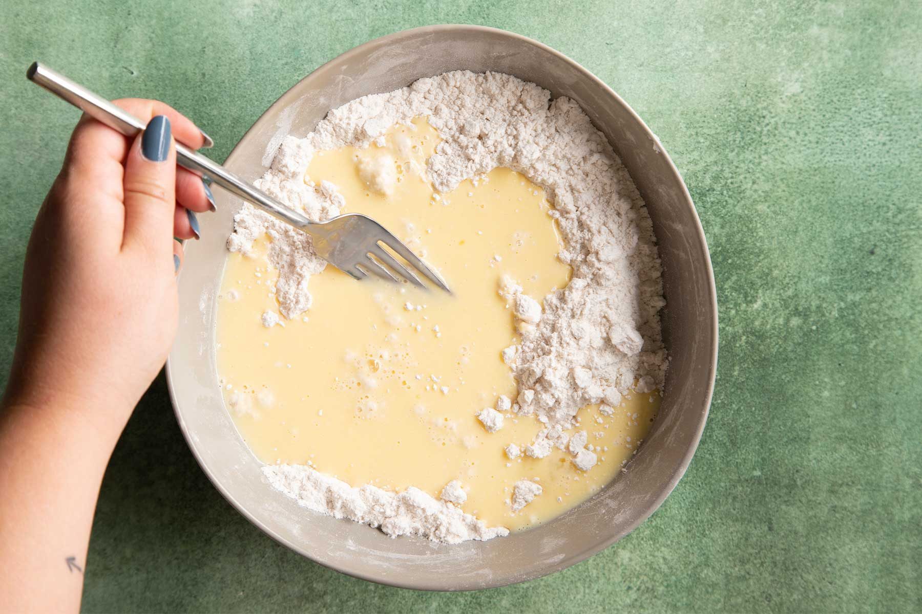 Overhead shot of mixing the egg mixture and flour mixture with fork in white bowl kept on top of a green marble countertop.