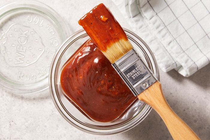 Top shot of barbeque sauce kept in a glass container with its lid beside it on top of a marble countertop with a check-pattern kitchen napkin kept beside it.