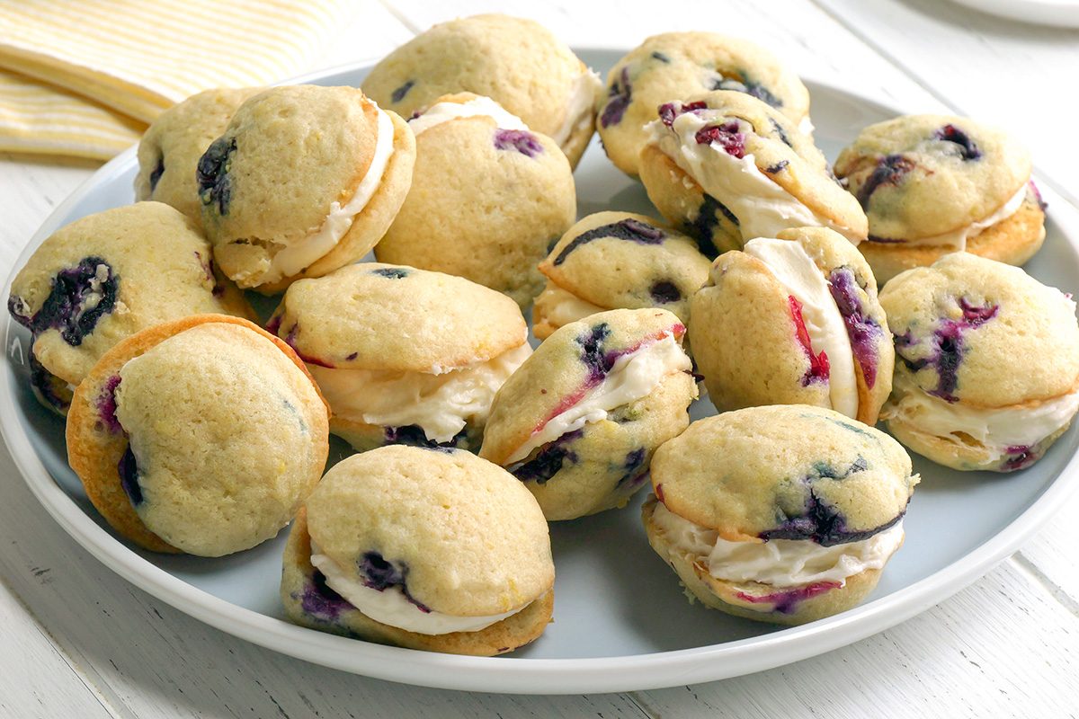 3/4th shot of Several lemon blueberry whoopie pies are arranged on a light blue plate, the plate rests on a white wooden surface, and in the blurred background, a yellow and white patterned cloth and the edges of white plates are visible