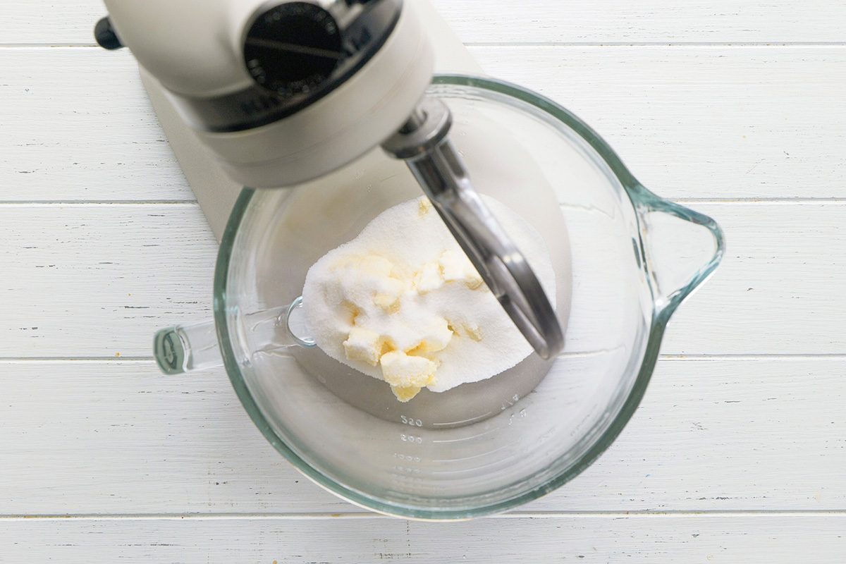 overhead shot of a clear glass mixing bowl attached to a white stand mixer; inside the bowl, there is a mixture of granulated white sugar and pale yellow butter; a flat beater attachment is submerged in the ingredients, the bowl and mixer are positioned on a white wooden surface